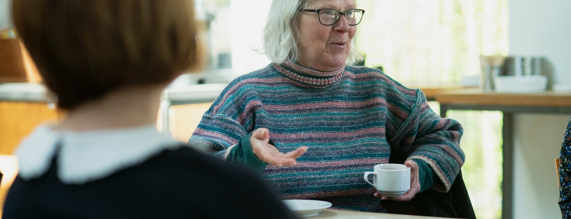 Bei einem Angehörigencafé erzählt eine Seniorin mit Brille, die eine Kaffeetasse in der Hand hält, den anderen Teilnehmerinnen von ihrem Alltag. 