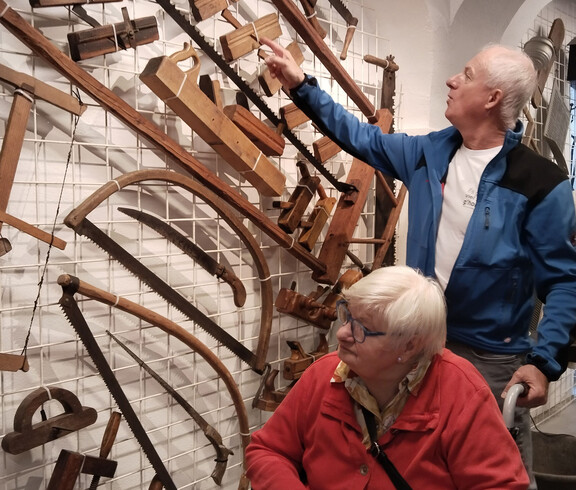Besucher*in im Rollstuhl betrachtet mit Begleiter alte Holzwerkzeuge an einer Ausstellungswand