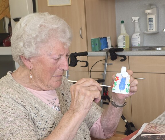 Eine Bewohnerin des Pflegewohnhauses gestaltet aufmerksam eine bunte Keramikfigur. Auf dem Tisch liegen Zeitungen als Unterlage sowie Pinsel und Farben. Die kreative Tätigkeit findet in einem der Gemeinschaftsräume statt.