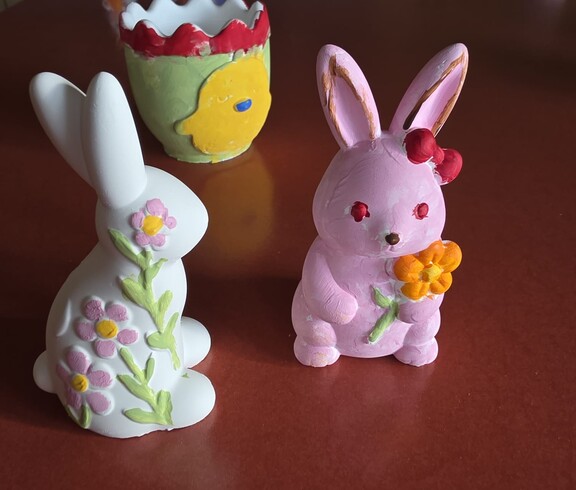 Farbenfrohe, handbemalte Osterfiguren aus Keramik – darunter zwei Hasen und ein dekorativer Blumentopf – stehen auf einem Tisch. Im Hintergrund sind weitere liebevoll gestaltete Osterdekorationen zu sehen.