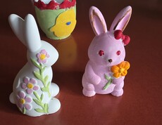 Farbenfrohe, handbemalte Osterfiguren aus Keramik – darunter zwei Hasen und ein dekorativer Blumentopf – stehen auf einem Tisch. Im Hintergrund sind weitere liebevoll gestaltete Osterdekorationen zu sehen.