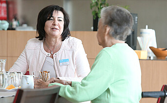 Eine ehrenamtliche Hospiz-Mitarbeiterin sitzt mit einer älteren Dame am Tisch bei der Kaffeejause, die beiden reden miteinander. 