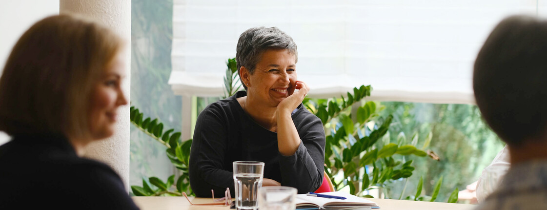 Eine lachende Frau sitzt mit anderen Frauen an einem Tisch und hört zu.