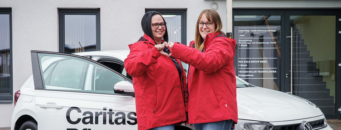 Zwei Caritas Mitarbeiterinnen stehen vor dem Caritas Auto der Mobilen Pflege und formen mit den Händen ein Herz.