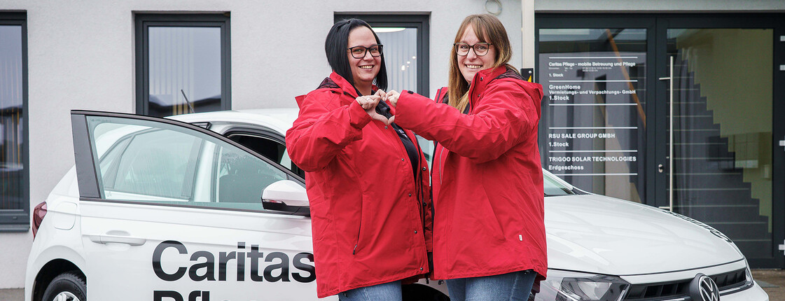 Zwei Caritas Mitarbeiterinnen stehen vor dem Caritas Auto der Mobilen Pflege und formen mit den Händen ein Herz.