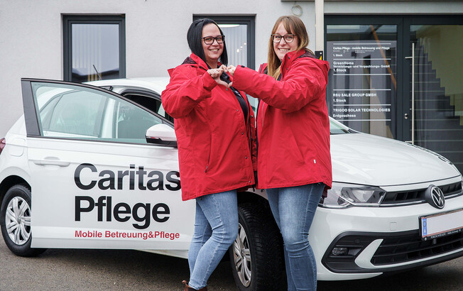 Zwei Caritas Mitarbeiterinnen stehen vor dem Caritas Auto der Mobilen Pflege und formen mit den Händen ein Herz.