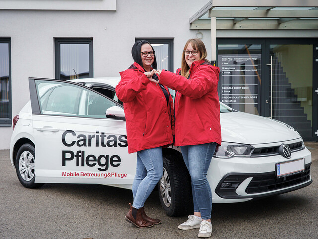 Zwei Caritas Mitarbeiterinnen stehen vor dem Caritas Auto der Mobilen Pflege und formen mit den Händen ein Herz.