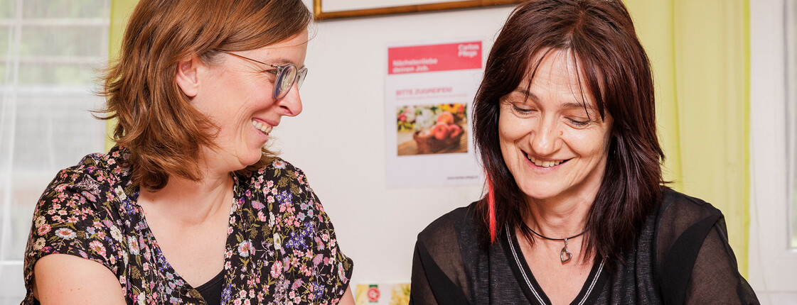 Zwei Mitarbeiterinnen der Caritas Pflege sitzen gemeinsam am Tisch und besprechen Unterlagen, die vor ihnen liegen.