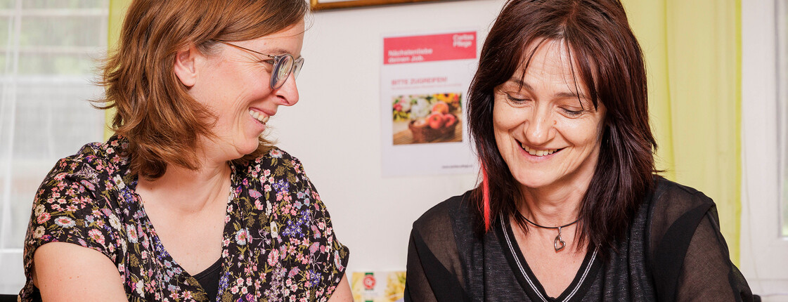 Zwei Mitarbeiterinnen der Caritas Pflege sitzen gemeinsam am Tisch und besprechen Unterlagen, die vor ihnen liegen.
