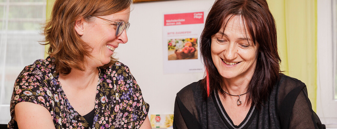 Zwei Mitarbeiterinnen der Caritas Pflege sitzen gemeinsam am Tisch und besprechen Unterlagen, die vor ihnen liegen.
