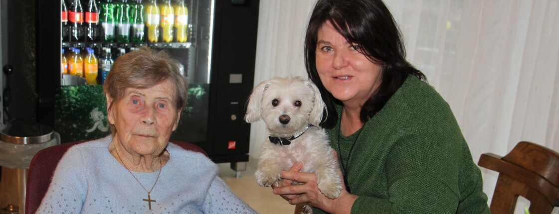 Eine Bewohnerin sitzt am Tisch vor ihrer Geburtstagstorte. Daneben sitzt eine Dame, die einen kleinen weißen Hund im Arm halten, und lächeln gemeinsam in die Kamera.