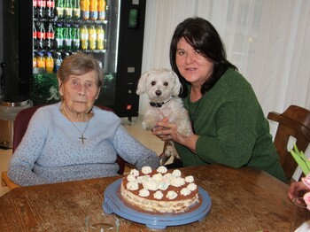 Eine Bewohnerin sitzt am Tisch vor ihrer Geburtstagstorte. Daneben sitzt eine Dame, die einen kleinen weißen Hund im Arm halten, und lächeln gemeinsam in die Kamera.