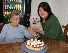 Eine Bewohnerin sitzt am Tisch vor ihrer Geburtstagstorte. Daneben sitzt eine Dame, die einen kleinen weißen Hund im Arm halten, und lächeln gemeinsam in die Kamera.