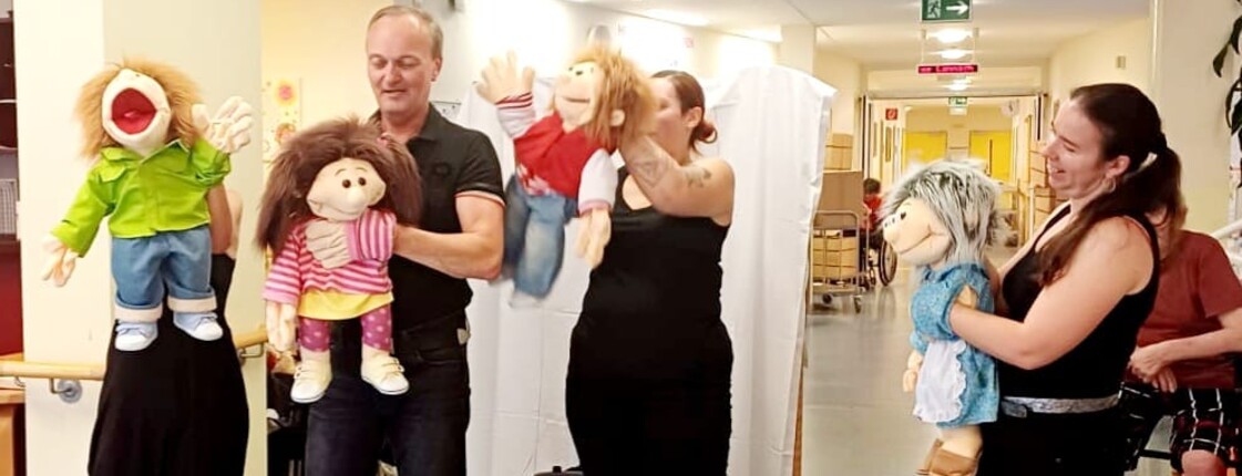 Vier schwarz gekleidete Puppenspieler stehen vor den Bewohner*innen des Caritas Pflegewohnhauses Lannach und führen eine Puppenshow auf. Die Szene zeigt eine lebendige Unterhaltung im Pflegealltag.