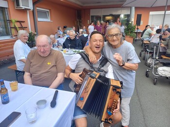 Eine Person mit einer Ziehharmonika sitzt zwischen den Gästen des Sommerfestes, während die Bewohnerinnen und Bewohner an festlich gedeckten Tischen im Innenhof zusammensitzen und der Musik lauschen.