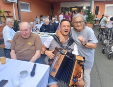 Eine Person mit einer Ziehharmonika sitzt zwischen den Gästen des Sommerfestes, während die Bewohnerinnen und Bewohner an festlich gedeckten Tischen im Innenhof zusammensitzen und der Musik lauschen.