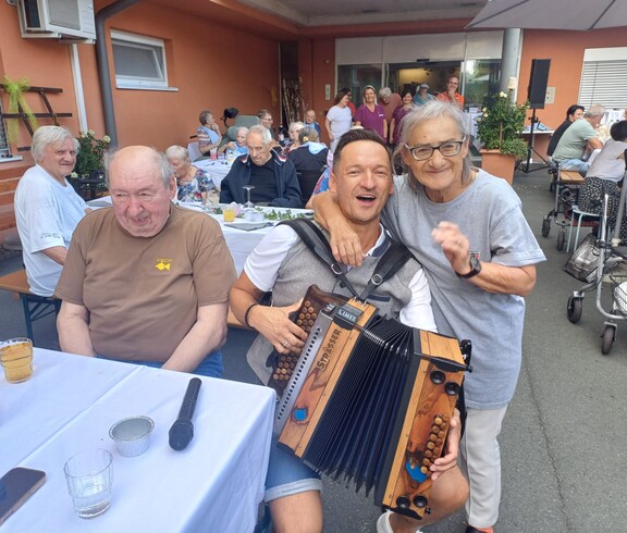 Eine Person mit einer Ziehharmonika sitzt zwischen den Gästen des Sommerfestes, während die Bewohnerinnen und Bewohner an festlich gedeckten Tischen im Innenhof zusammensitzen und der Musik lauschen.