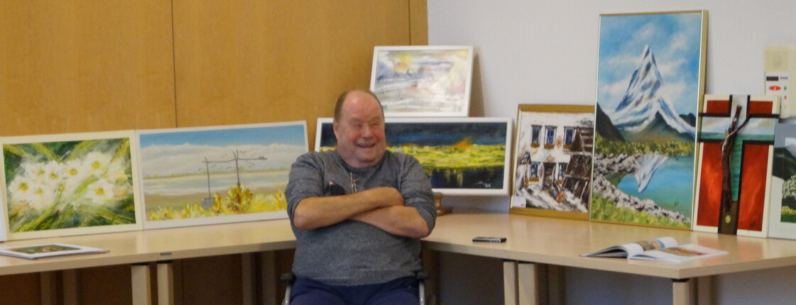 Bilder stehen im Hintergrund auf einem Tisch und lehnen an der Wand. Davor im Rollstuhl sitzt ein Mann mit den Armen verschränkt vor der Brust.