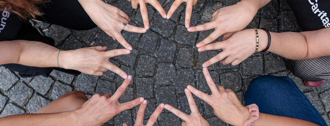 Fünf Personen sitzen im Kreis in der Hocke und formen mit den Zeige- und Mittelfingern einen Stern.