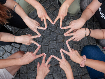 Fünf Personen sitzen im Kreis in der Hocke und formen mit den Zeige- und Mittelfingern einen Stern.