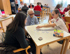 Jugendliche und Seniorin sitzen gemeinsam am Tisch und spielen Memory im hellen Festsaal.