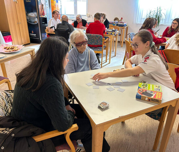 Jugendliche und Seniorin sitzen gemeinsam am Tisch und spielen Memory im hellen Festsaal.