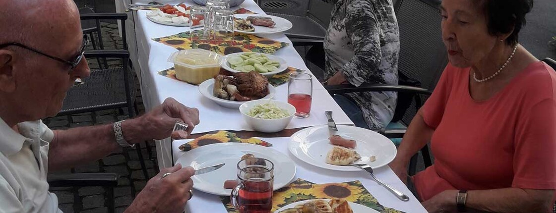 Zwei Frauen und ein Mann sitzen an einer gedeckten Tafel und essen Gegrilltes.