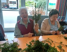 Bewohnerinnen des Pflegewohnhauses binden gemeinsam Palmbuschen und sitzen dabei in einem hellen Gemeinschaftsraum. Auf dem Tisch liegen frische Zweige bereit, während in entspannter Atmosphäre gearbeitet und geplaudert wird.