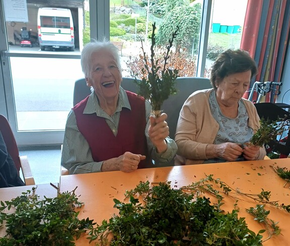 Bewohnerinnen des Pflegewohnhauses binden gemeinsam Palmbuschen und sitzen dabei in einem hellen Gemeinschaftsraum. Auf dem Tisch liegen frische Zweige bereit, während in entspannter Atmosphäre gearbeitet und geplaudert wird.