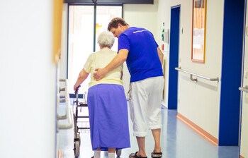 Ein Pfleger stützt eine ältere Dame beim Gehen mit ihrem Rollator.