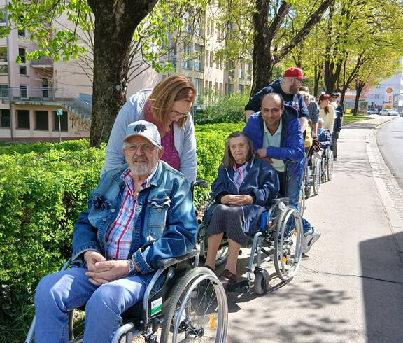Mehrere Menschen im Rollstuhl werden von Begleitpersonen entlang eines grünen Gehwegs durch die Stadt geschoben.