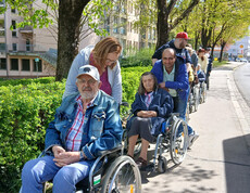 Mehrere Menschen im Rollstuhl werden von Begleitpersonen entlang eines grünen Gehwegs durch die Stadt geschoben.