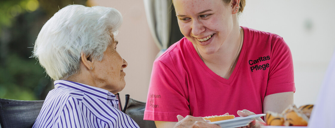 Eine Pflegerin reicht einer Dame einen Teller mit Kuchen, während sie sich gegenseitig anlächeln.