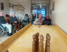 Bewohnerin stößt eine Kugel über die Holzbahn, andere sehen im Aufenthaltsraum zu.