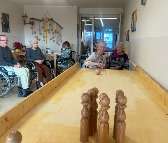 Bewohnerin stößt eine Kugel über die Holzbahn, andere sehen im Aufenthaltsraum zu.