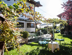 Im Garten eines Gasthauses stehen Stühle und Tische unter einer Weinlaufe. Auf dem Tisch stehen eine Flasche und ein Glas Rotwein.