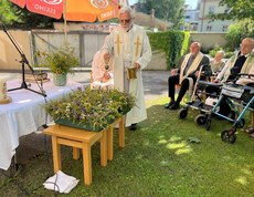 Priester segnet Kräutersträuße im Garten, mehrere Senior*innen sitzen im Halbkreis