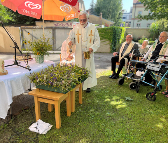 Priester segnet Kräutersträuße im Garten, mehrere Senior*innen sitzen im Halbkreis