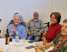 Gruppe älterer Frauen und Besucher*innen sitzen fröhlich an einem Tisch mit Getränken