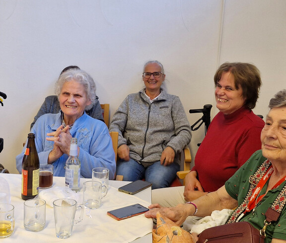 Gruppe älterer Frauen und Besucher*innen sitzen fröhlich an einem Tisch mit Getränken