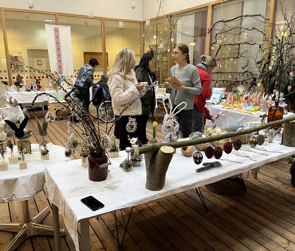Tische mit handgefertigter Osterdekoration bei einem Basar im Innenbereich eines Pflegewohnhauses, mehrere Personen betrachten die Ausstellungsstücke