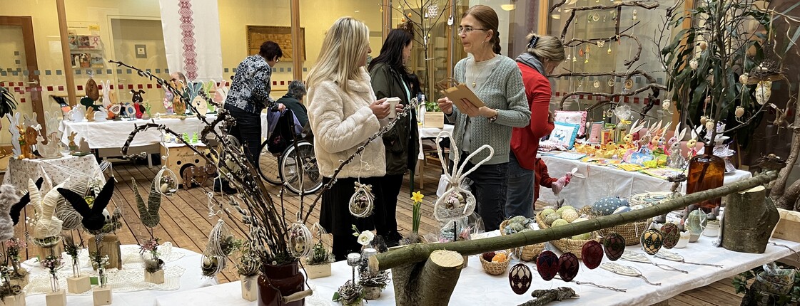 Tische mit handgefertigter Osterdekoration bei einem Basar im Innenbereich eines Pflegewohnhauses, mehrere Personen betrachten die Ausstellungsstücke