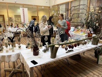 Tische mit handgefertigter Osterdekoration bei einem Basar im Innenbereich eines Pflegewohnhauses, mehrere Personen betrachten die Ausstellungsstücke