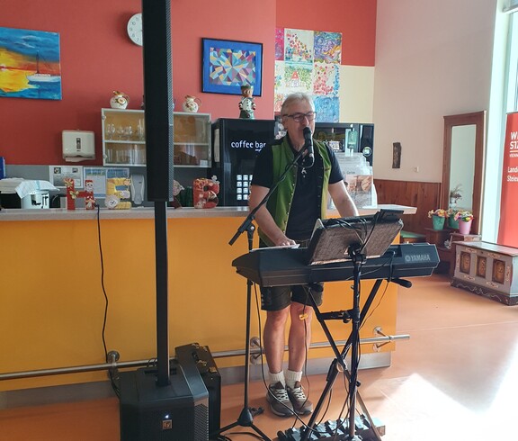 Eine Person steht an einem Keyboard mit Mikrofon und Lautsprecheranlage und sorgt in einem hellen Aufenthaltsraum mit farbenfrohen Wandbildern und Kunstwerken für die musikalische Unterhaltung der Veranstaltung.