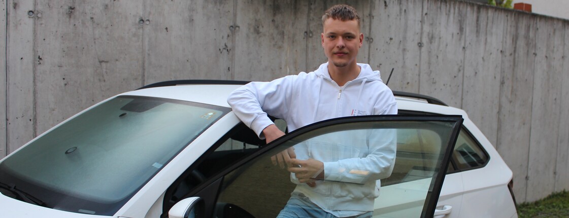 Junger Mann steht hinter offener Fahrertüre eines Autos.
