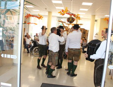 Mehrere Mitglieder der Pischelsdorfer Schuhplattler in traditioneller Tracht führen im festlich mit Herbstdekoration geschmückten Aufenthaltsbereich des Caritas Pflegewohnhauses einen Tanz auf, während Bewohnerinnen und Bewohner das Geschehen von ihren Sitzplätzen aus verfolgen.