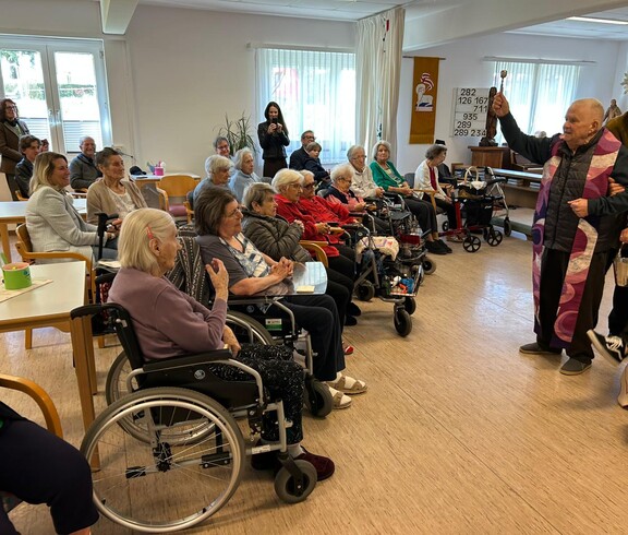 Geistlicher weiht die mitgebrachten Speisen vor sitzenden Bewohner*innen im Gemeinschaftsraum.