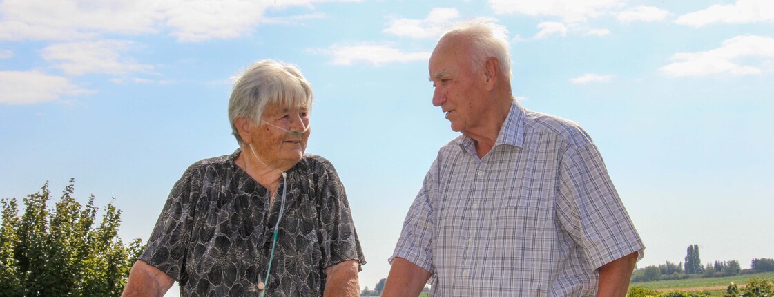 Zwei Personen aus dem Haus Lisa steht mit Rollatoren im Freien und unterhält sich. Die Frau hat eine Sauerstoffnasenbrille, und beide genießen den sonnigen Tag in einer ländlichen Umgebung.