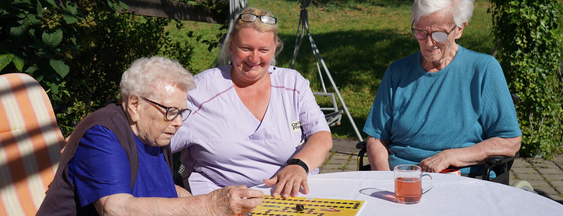 Spiele spielen im Haus St. Martin Zwei Bewohnerinnen sitzen zusammen mit einer Pflegekraft draußen an einem Tisch und spielen ein Brettspiel. Alle drei wirken konzentriert und genießen die Aktivität im Freien, während sie bei sonnigem Wetter beisammen sind.