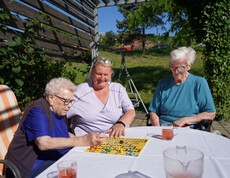 Zwei Bewohnerinnen sitzen zusammen mit einer Pflegekraft draußen an einem Tisch und spielen ein Brettspiel. Alle drei wirken konzentriert und genießen die Aktivität im Freien, während sie bei sonnigem Wetter beisammen sind.